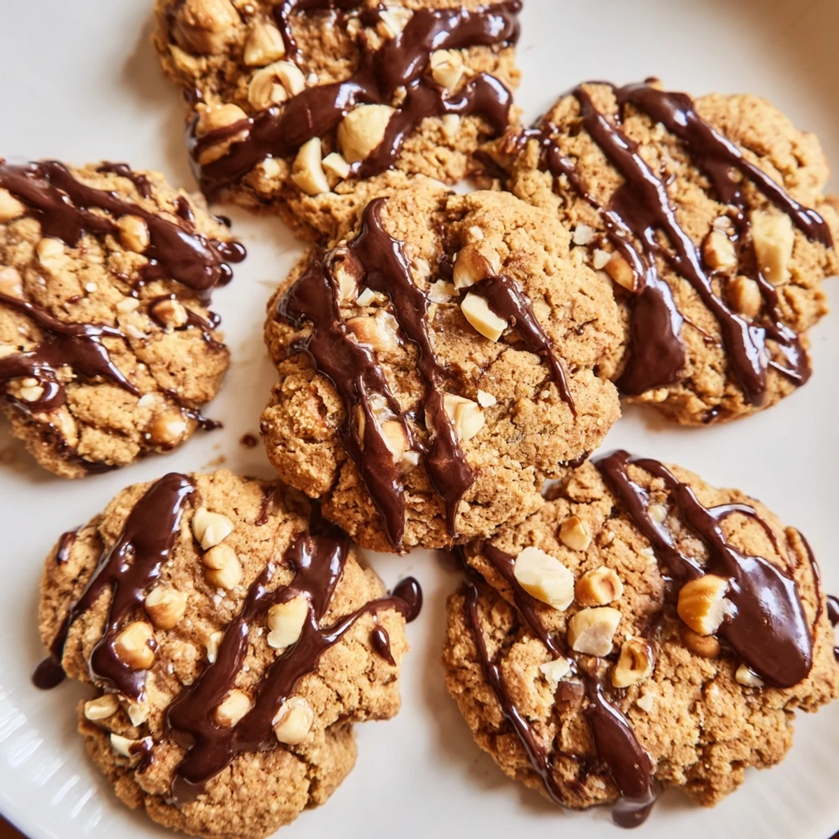 Freshly baked Holiday Glow Hazelnut Drizzle Vegan Cookies are stacked on a wooden board with toasted hazelnuts.