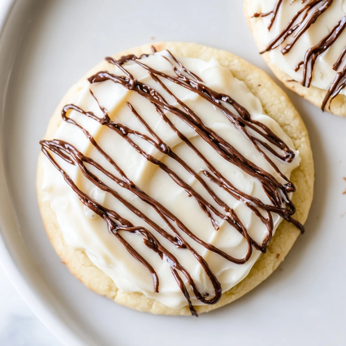 Soft, buttery sugar cookies topped with creamy vanilla frosting and a rich hazelnut chocolate drizzle, arranged on a white plate with a cup of coffee.